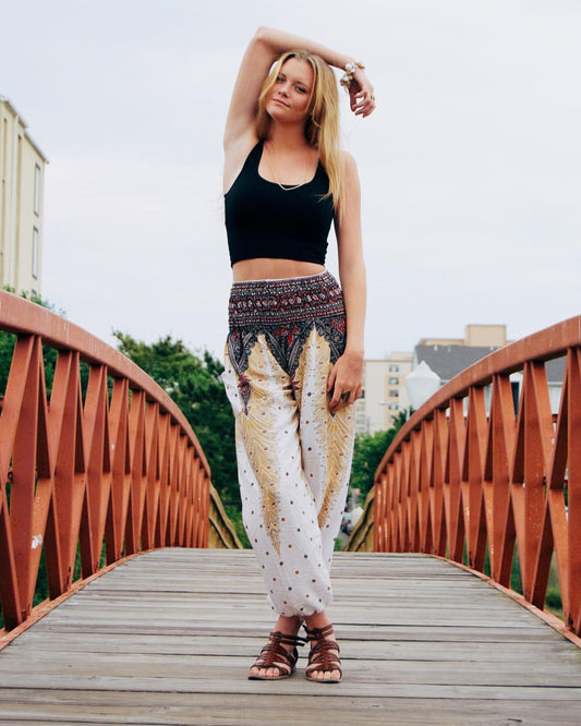Woman posing on a wooden bridge wearing a black top and white Thai harem yoga pants with peacock print