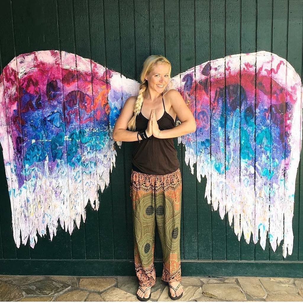 Woman wearing olive green Thai harem yoga pants with mandala print and black top standing between colorful angel wings against a dark green wall.