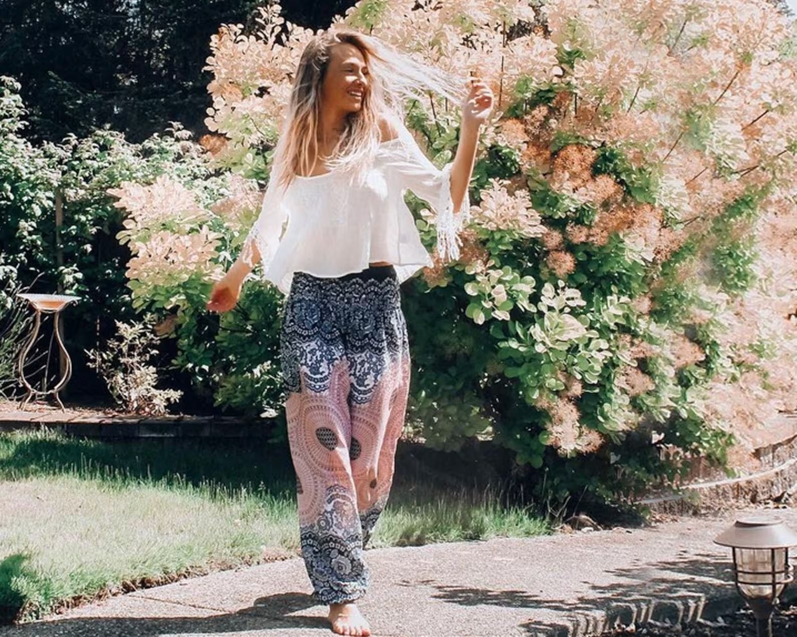 Woman walking in a garden with flowers and trees in the background wearing gray Thai harem yoga pants with mandala print and white top