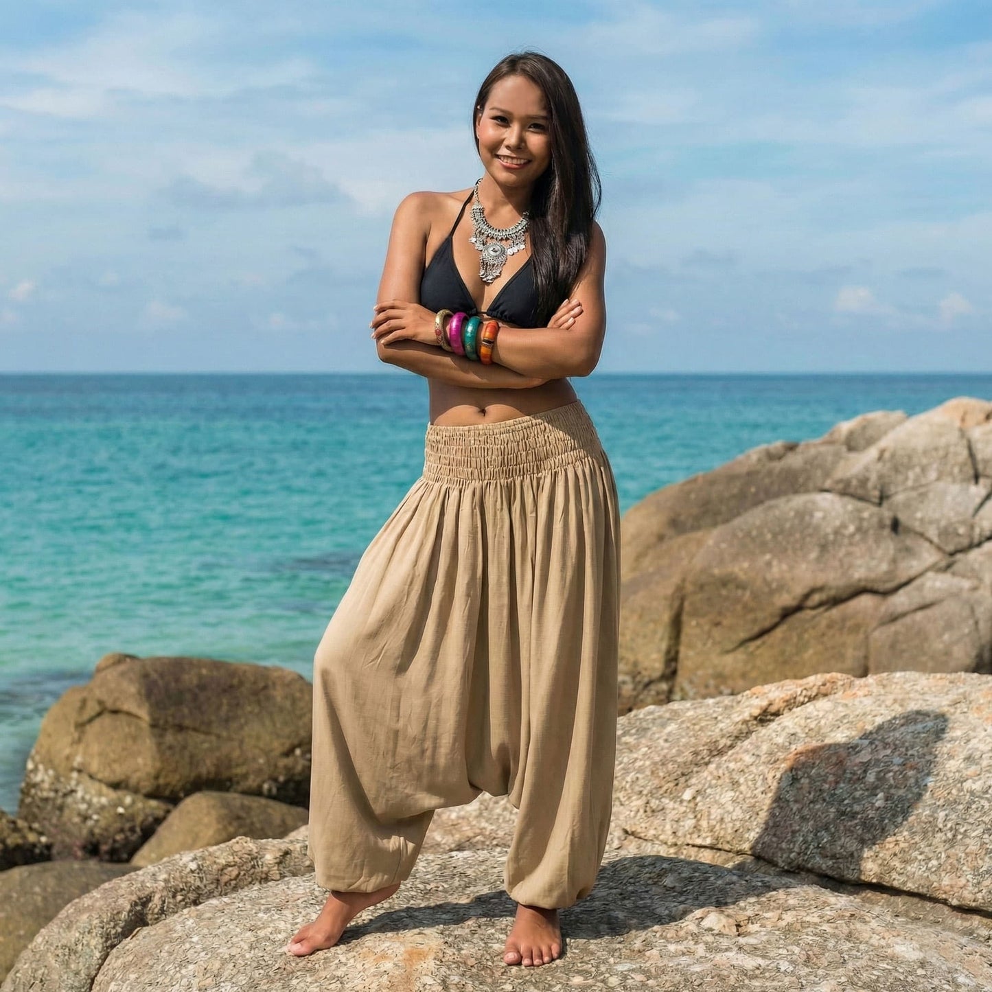 Woman standing on rocks by the ocean wearing a black bikini top and beige jumpsuit thai harem pants.