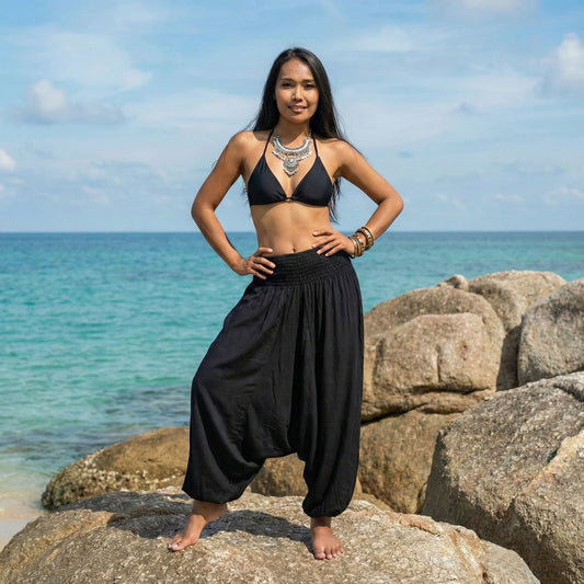 Woman in black bikini top and black thai harem pants jumpsuit standing on rocks by the ocean