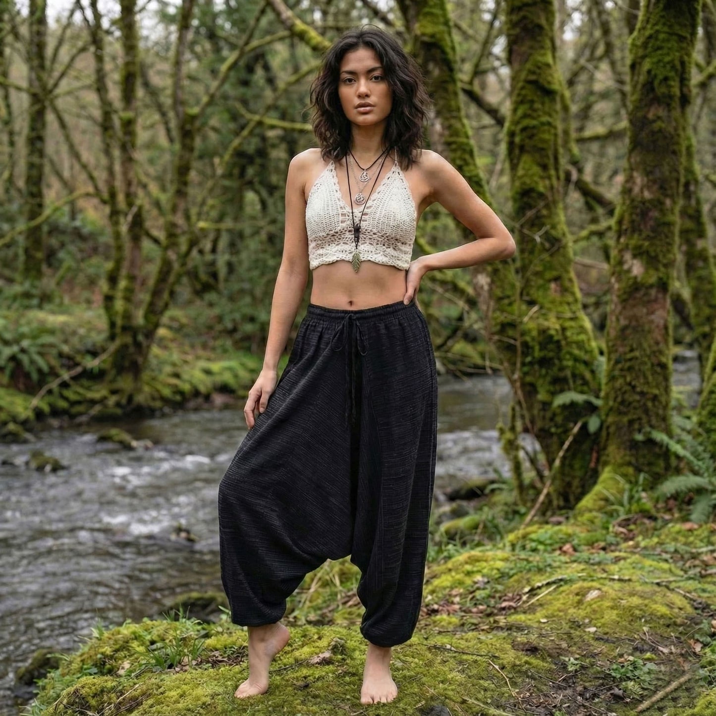 Woman standing in a forest with a stream in the background wearing black pinstripe thai harem pants and white top