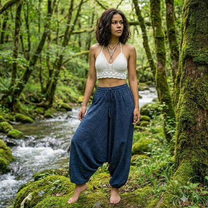 Woman in a white top and blue pinstripe thai harem pants standing in a lush green forest with a stream.