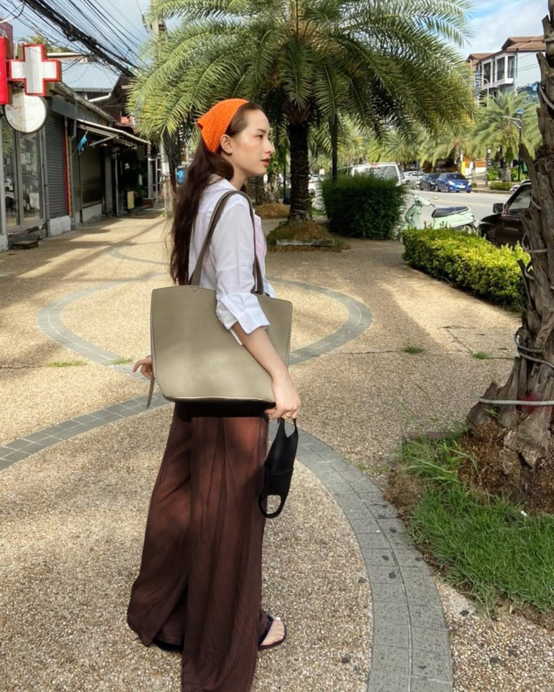 Woman in brown thai wrap pants skirt and white top walking outdoors on a sidewalk with palm trees and buildings in the background