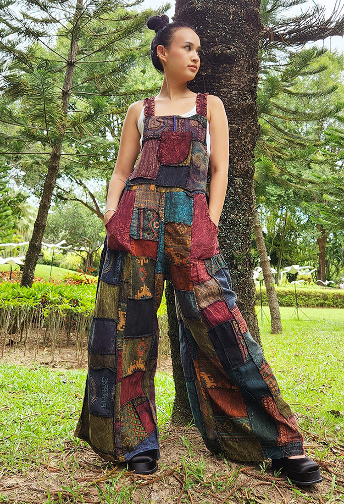 Female model wearing brown patchwork overall jumpsuit in a park, front view.