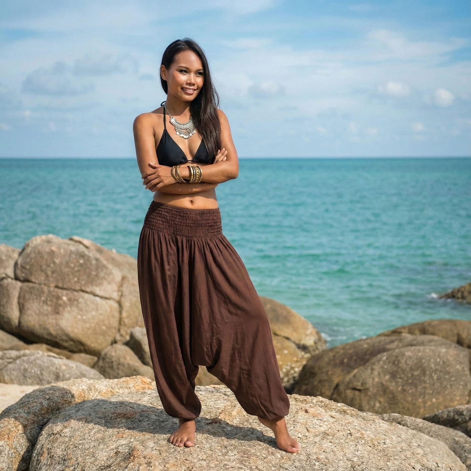Woman standing on rocks by the ocean with a blue sky waering a brown 2-in-1 jumpsuit harem pants and black bikini top