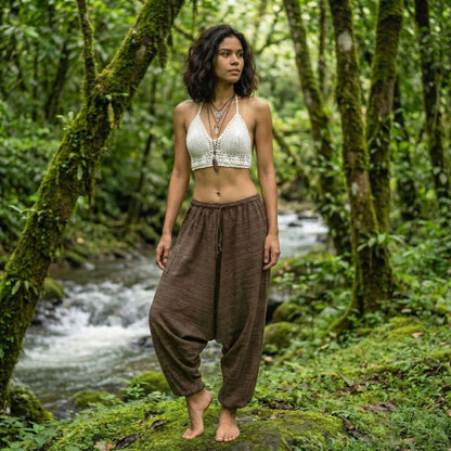Woman in a white top and brown pinstripe thai harem pants standing in a lush green forest with a stream.