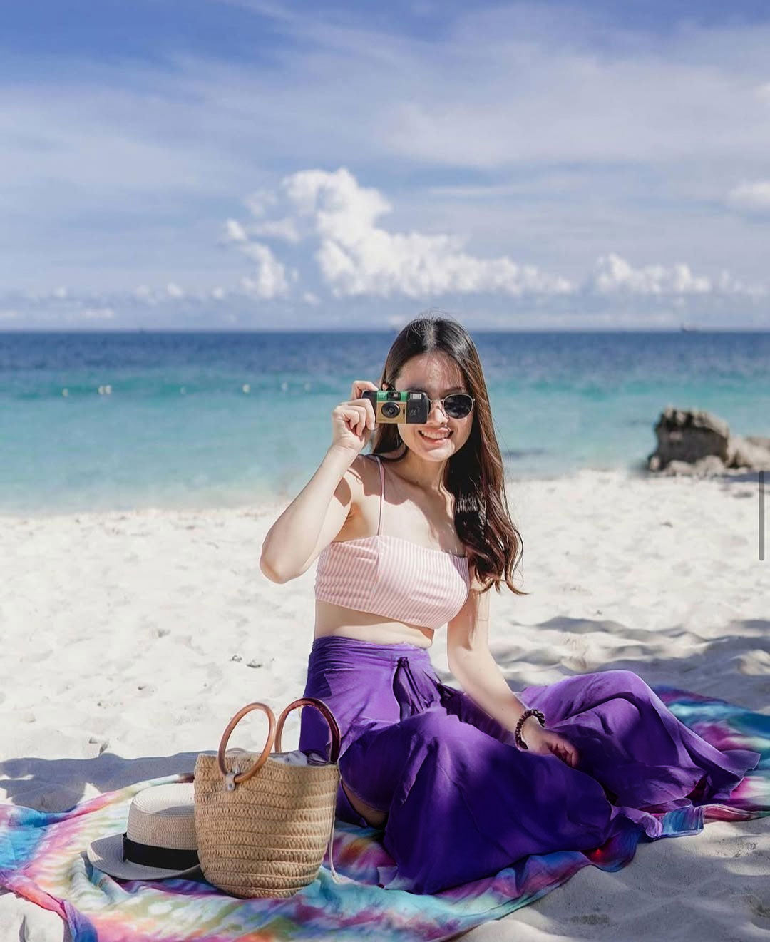 Woman sitting on a beach with a colorful blanket, holding sunglasses, and wearing a pink top and purple Thai Wrap Pants skirt.
