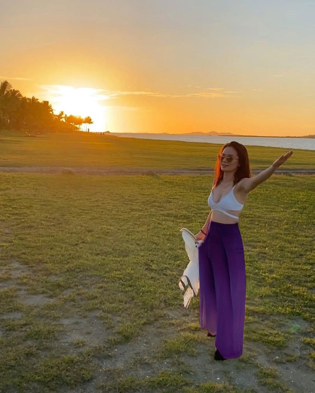 Woman in a white top and purple Thai wrap pants standing in a field with a sunset in the background