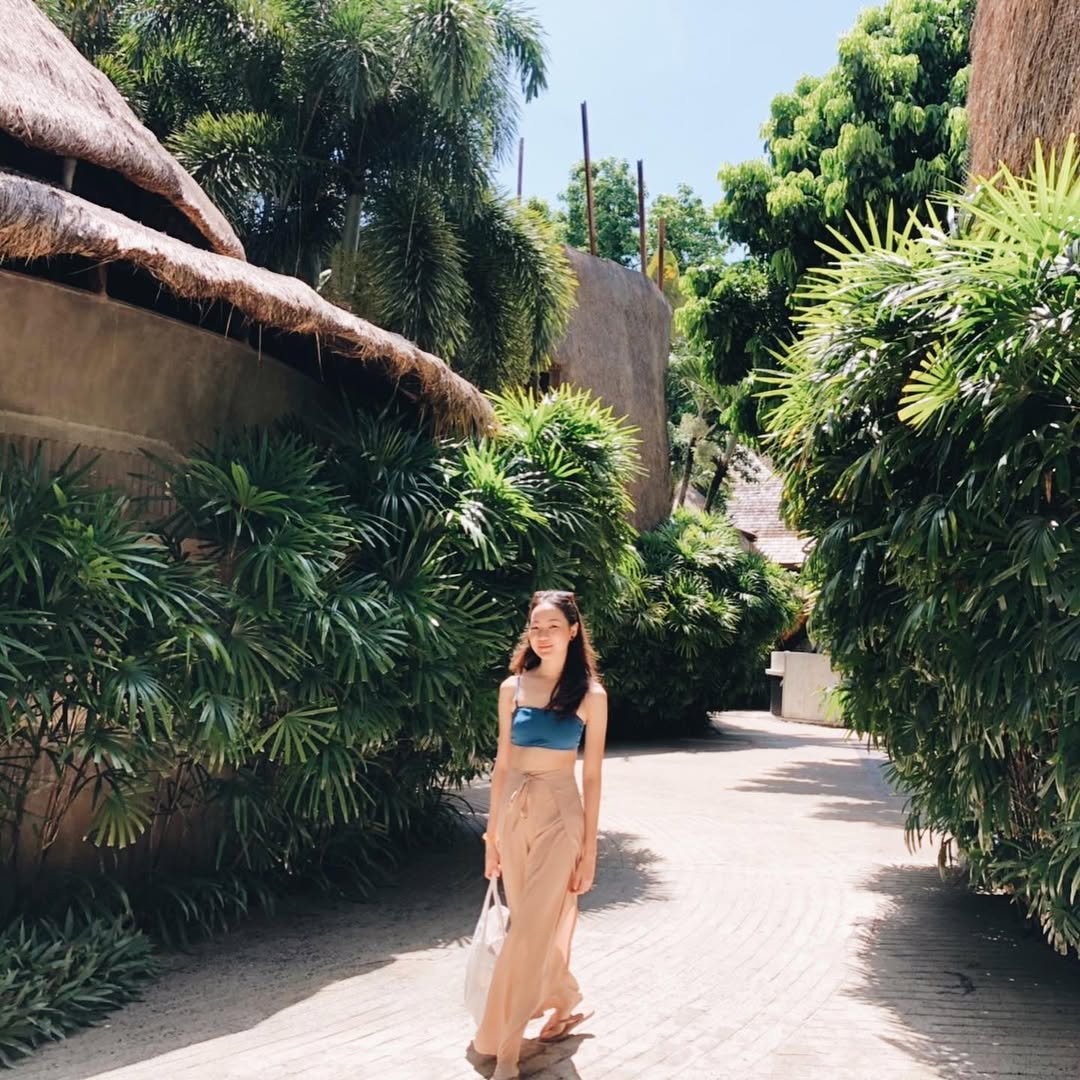 Woman in beige Thai wrap pants skirt with green top walking along a path with tropical plants and thatched-roof structures.