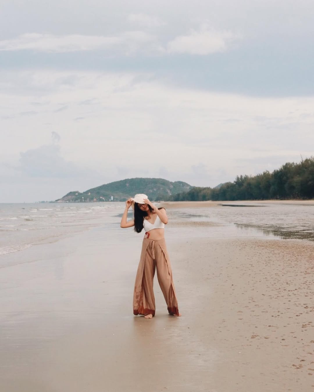 Woman in beige Thai wrap pants skirt with white top standing on a beach with mountains in the background