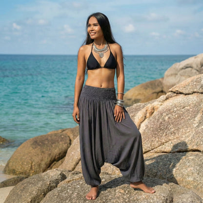 Woman in black bikini top and gray thai jumpsuit harem pants standing on rocks by the ocean.