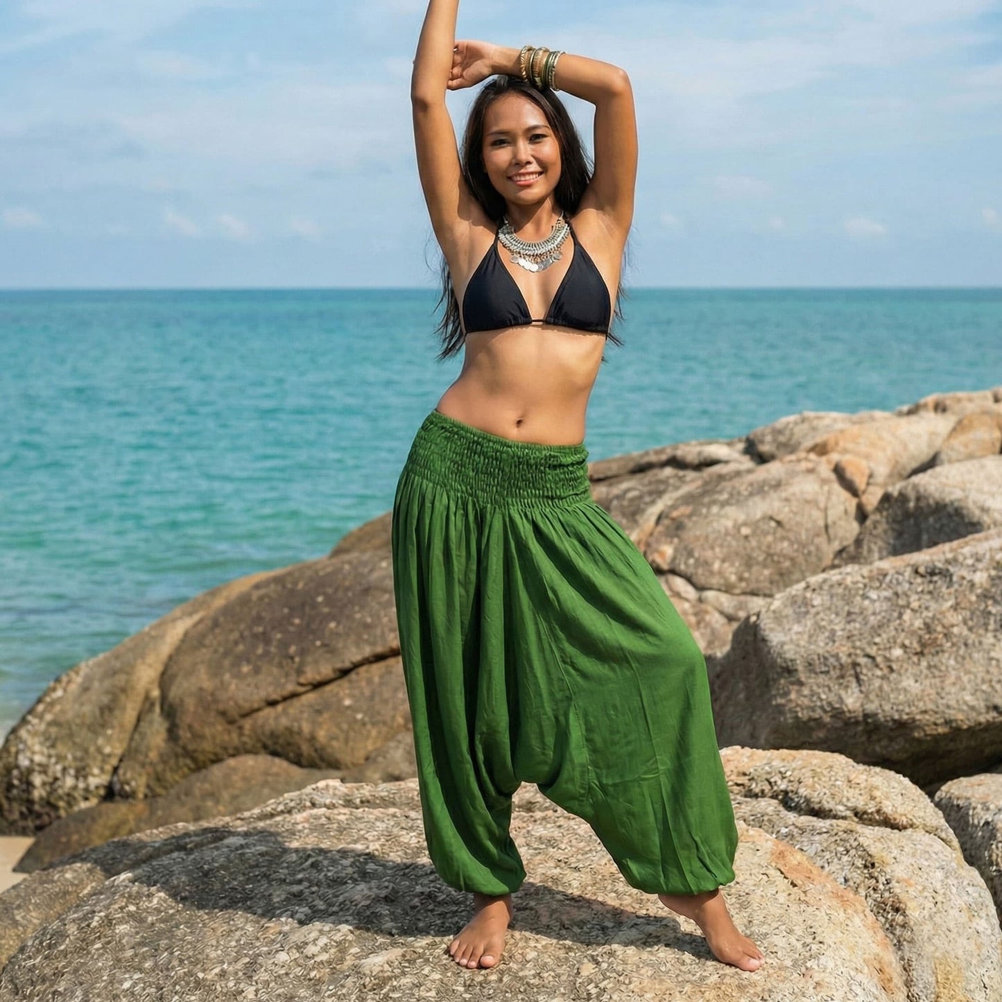 Woman in black bikini top and green jumpsuit thai harem pants standing on rocks by the ocean.