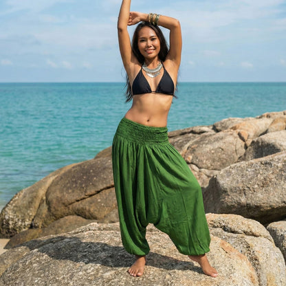 Woman in black bikini top and green jumpsuit thai harem pants standing on rocks by the ocean.