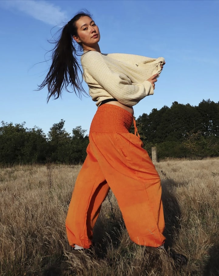 Person in beige sweater and orange my thai harem pants standing in a field with trees in the background
