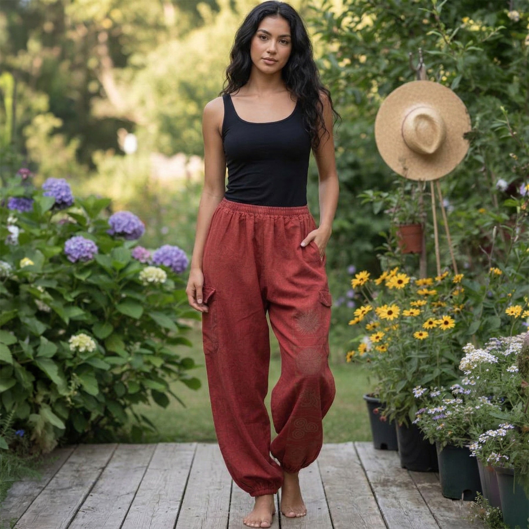 Woman in black tank top and red boho harem pants standing in a garden with flowers and a straw hat in the background.