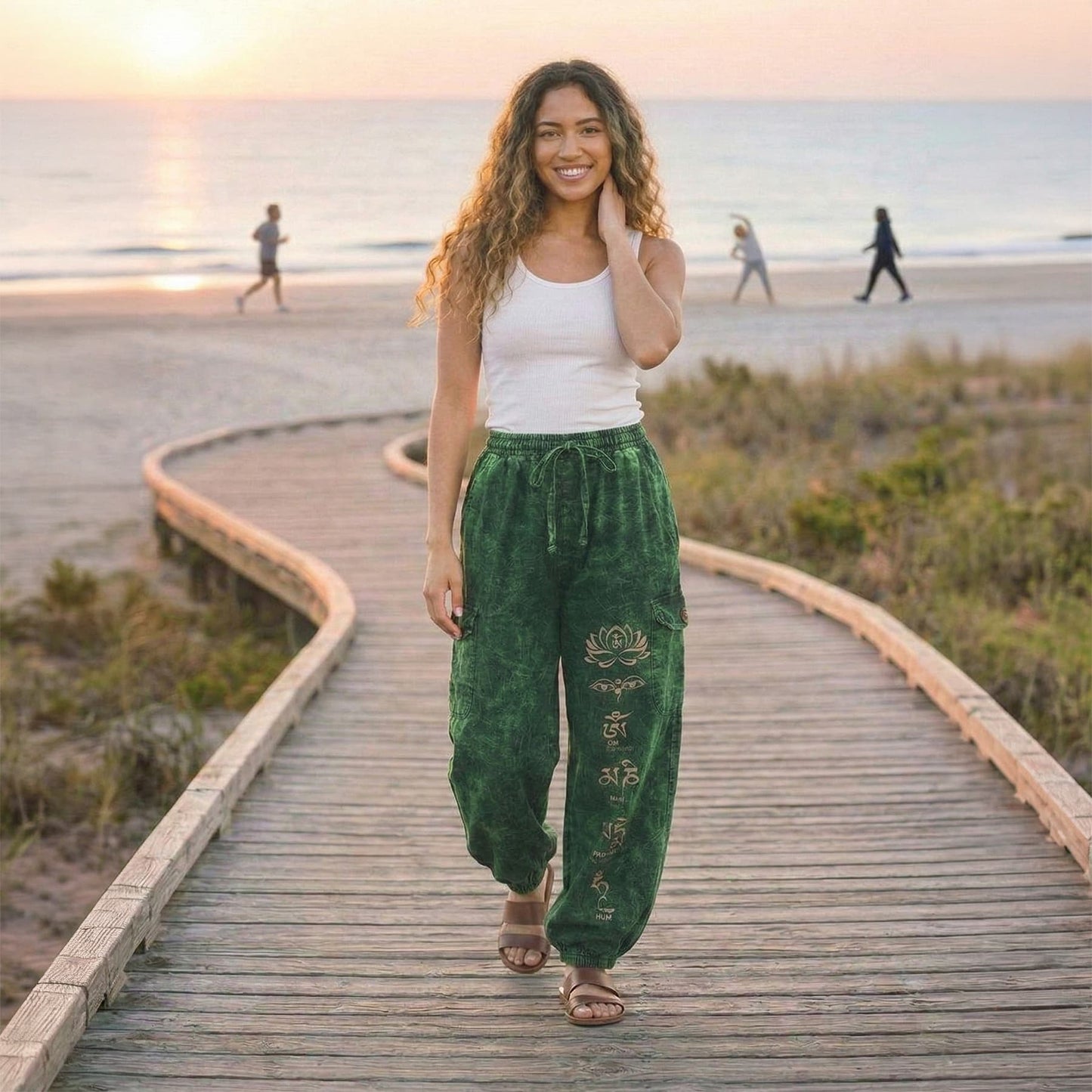 Female model wearing green boho lotus festival harem pants on a wooden path at the beach, front view.