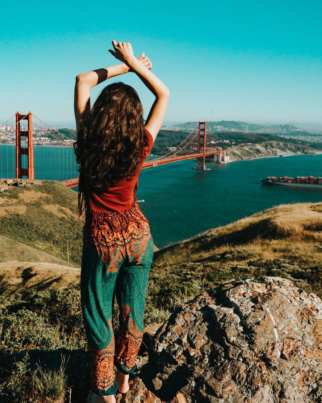 Woman wearing turquoise Thai harem yoga pants with mandala print and red top standing on a hill overlooking the Golden Gate Bridge with arms raised.