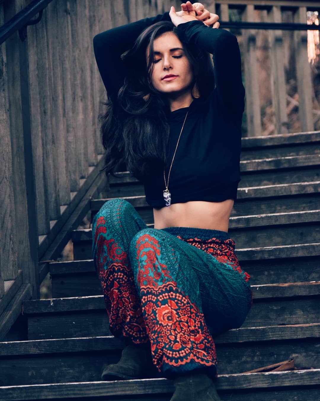 Woman sitting on outdoor wooden steps wearing black crop top and turquoise Thai harem yoga pants with mandala print