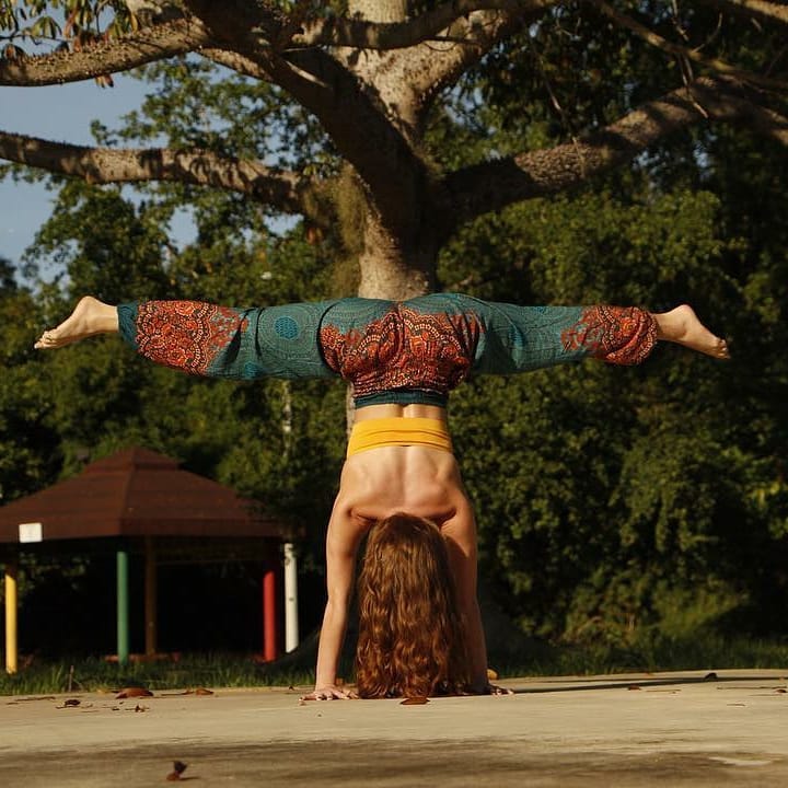 Woman doing yoga handstand in park wearing turquoise Thai harem yoga pants with mandala print and orange top