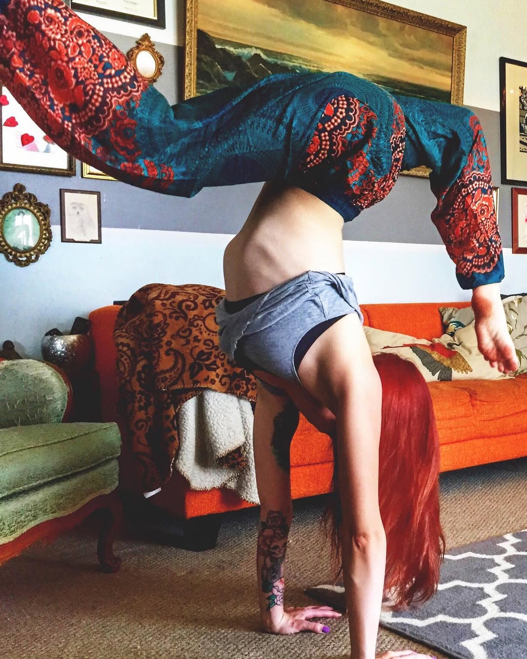 Woman wearing turquoise Thai harem yoga pants with mandala print and grey top doing a handstand in a room with colorful decor and furniture.