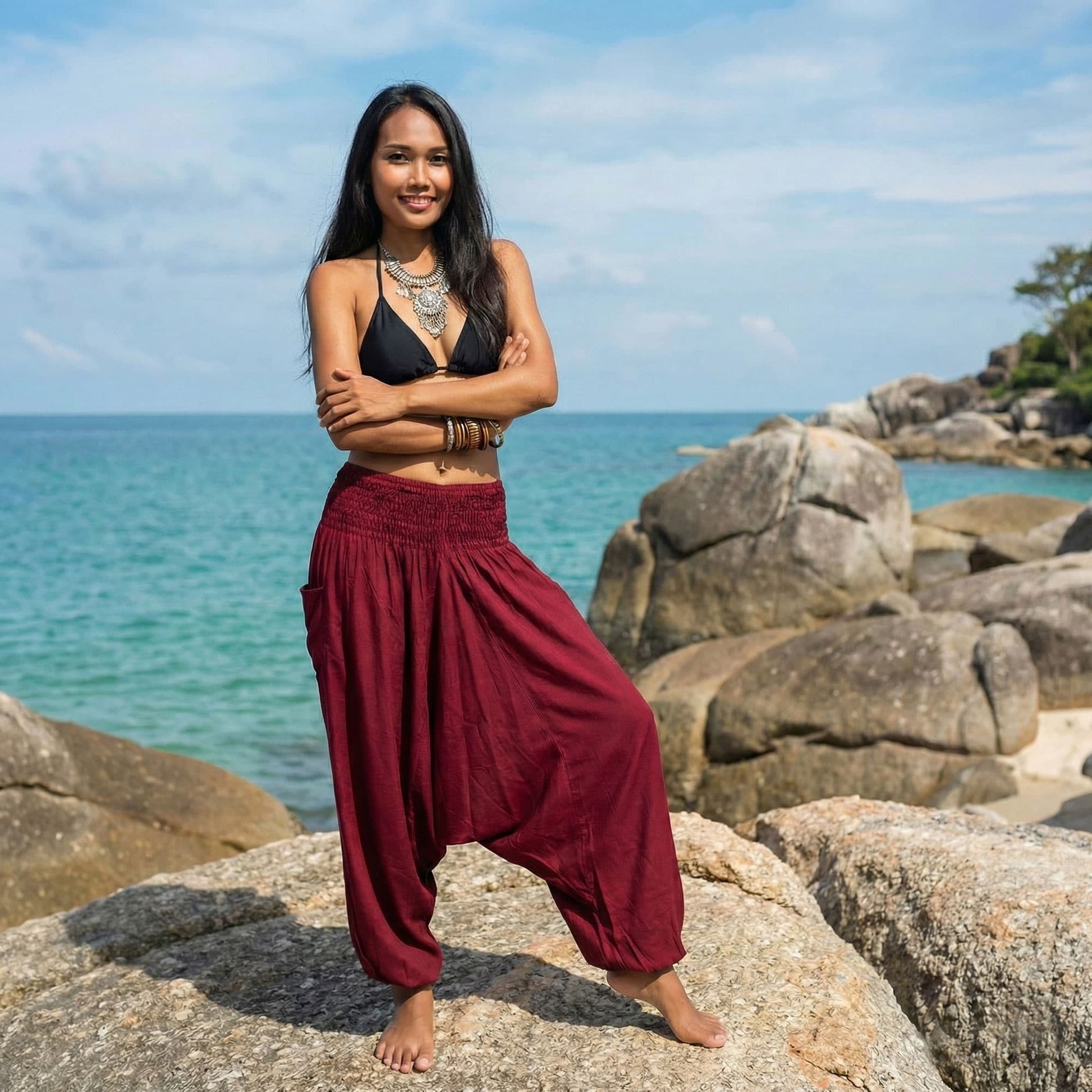 Woman standing on rocks by the ocean wearing a black top and maroon jumpsuit thai harem pants.