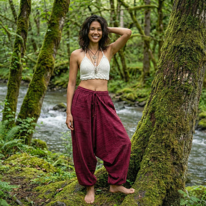 Woman standing in a forest by a stream wearing a white top and maroon pinstripe thai harem pants.
