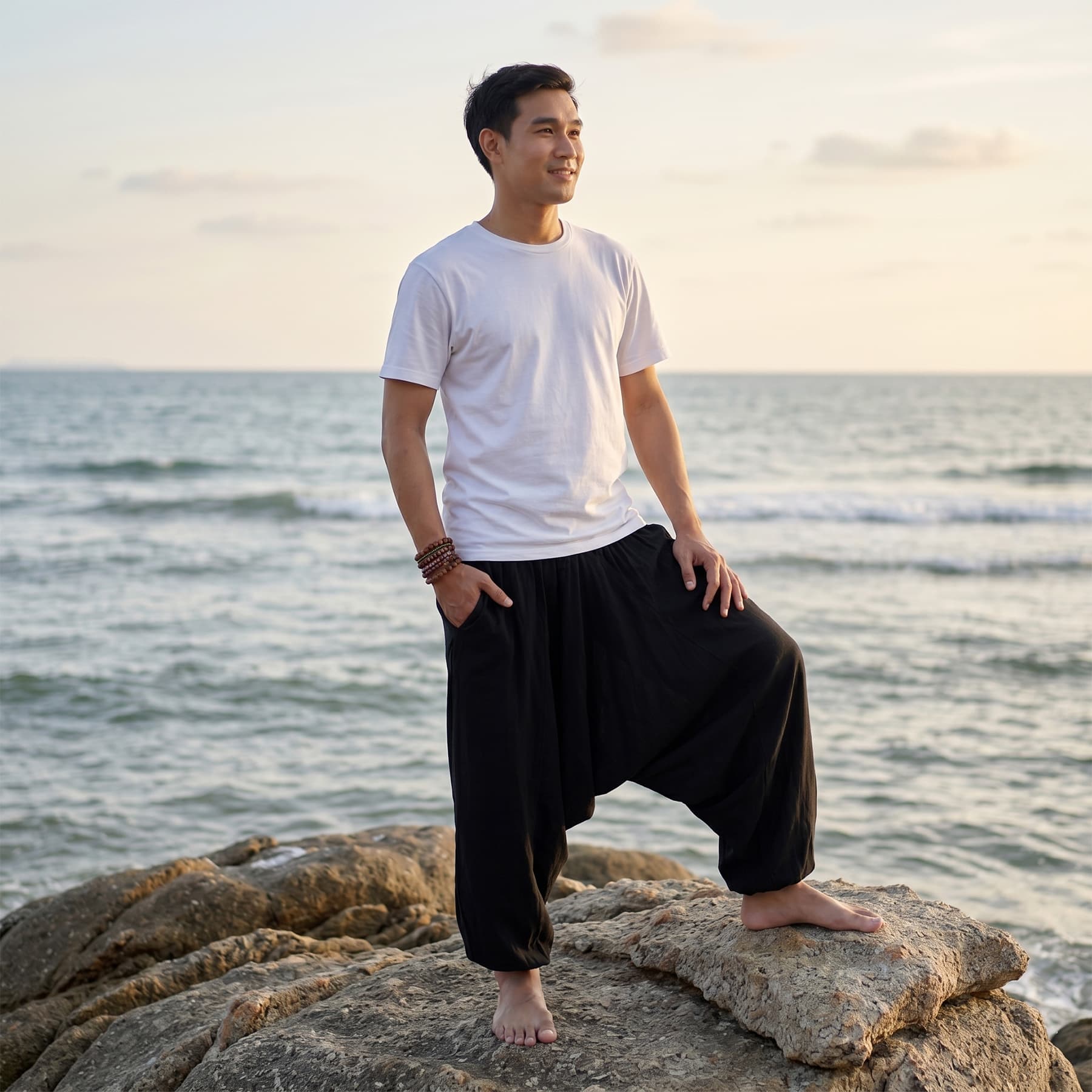 Male model on rocky shores wearing solid black Men's drop crotch harem pants under the blue sky.