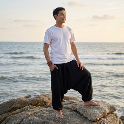 Male model on rocky shores wearing solid black Men's drop crotch harem pants under the blue sky.