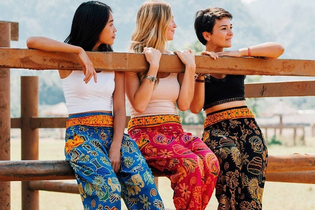 Three women wearing colorful patterned harem pants and matching tank tops lean against a rustic wooden fence in an outdoor setting.