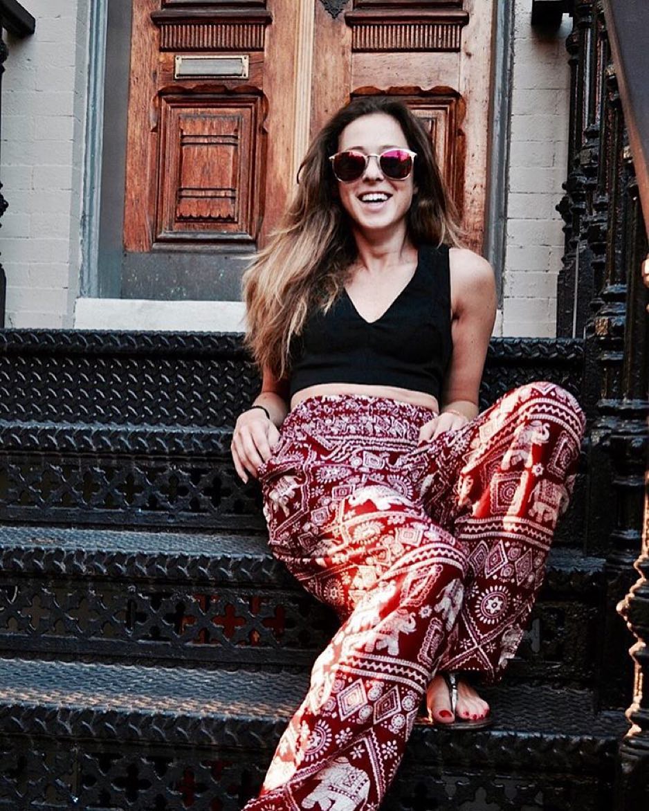 Woman sitting on steps wearing a black crop top and red Thai Elephant Harem Yoga Pants, with a wooden door in the background.