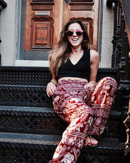 Woman sitting on steps wearing a black crop top and red Thai Elephant Harem Yoga Pants, with a wooden door in the background.
