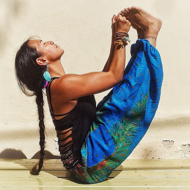 Person in a yoga pose wearing a blue Thai harem yoga pants with peacock print and black top against a light background