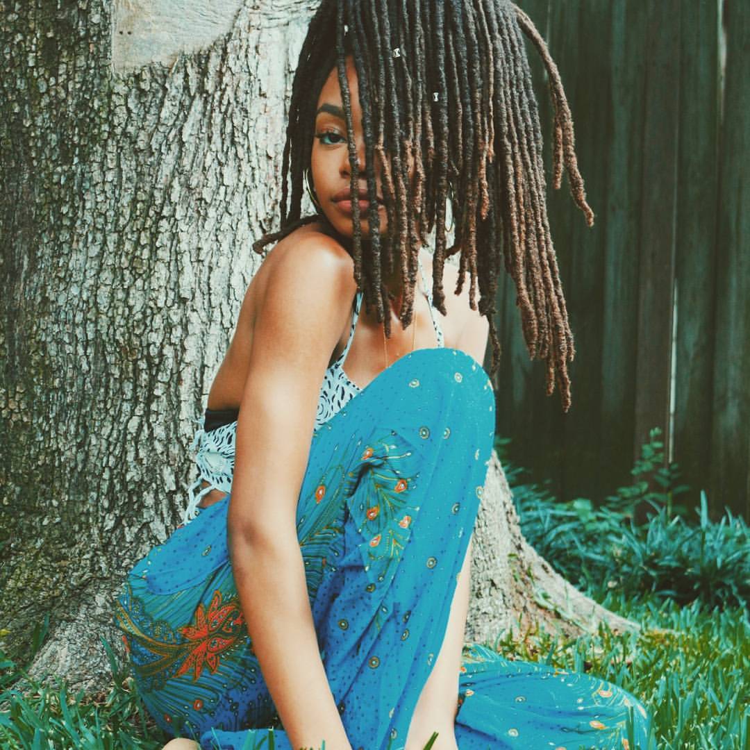 Woman blue Thai harem yoga pants with peacock print and  white top sitting against a tree in a forest setting