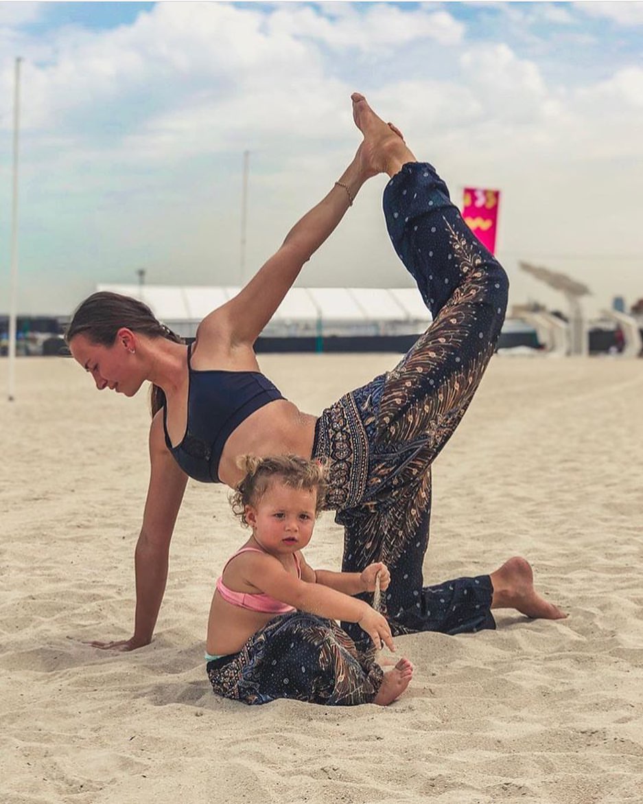 Woman and child performing a yoga pose on a sandy beach with a cloudy   sky both wearing green Thai harem yoga pants with peacock print and black top