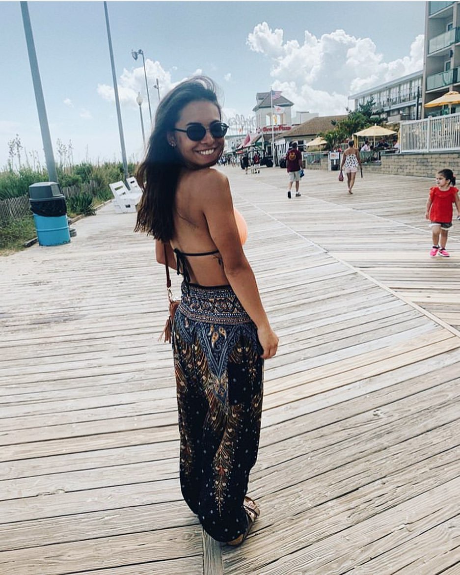 Woman in green Thai harem yoga pants with peacock print and orange top and sunglasses on a wooden boardwalk with people in the background.