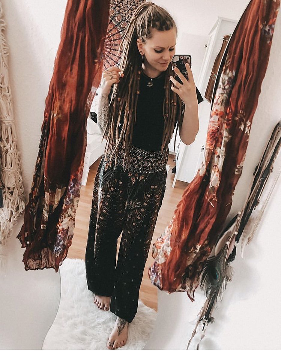 Woman taking a mirror selfie wearing a green Thai harem yoga pants with peacock print and black top standing in a room with decorative elements.