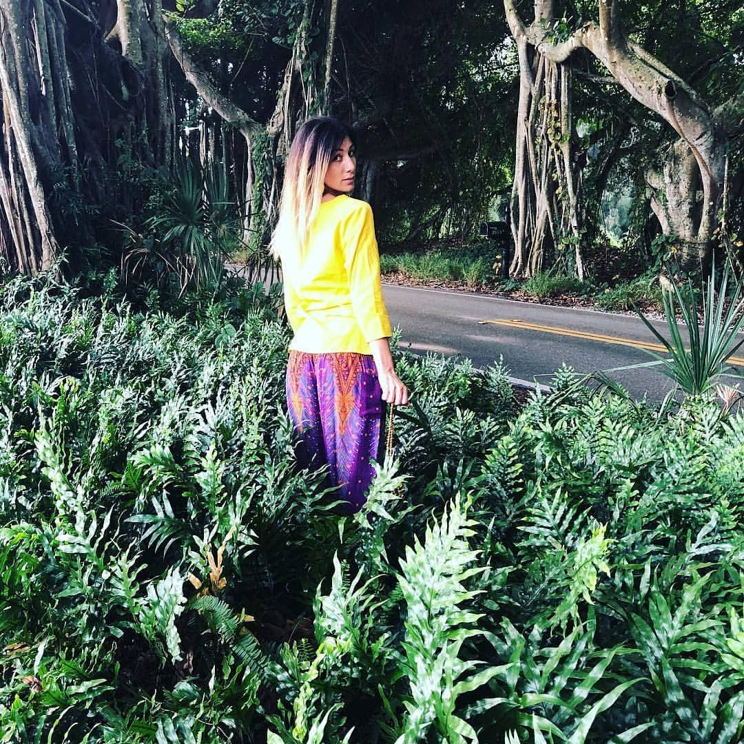 Woman in yellow shirt and a purple Thai harem yoga pants with peacock print standing among green plants with a road and trees in the background