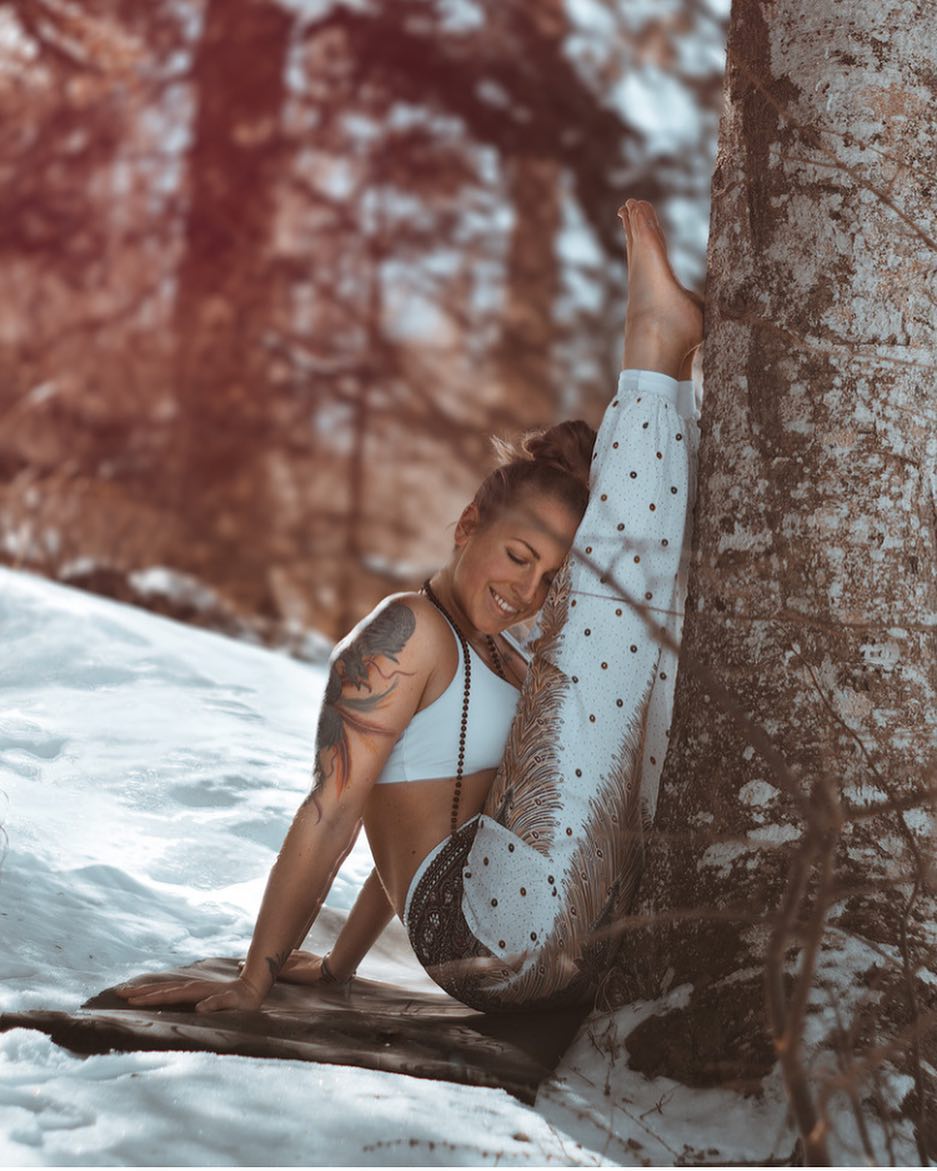 Person in white Thai harem yoga pants with peacock print and white top leaning against a tree in a snowy forest