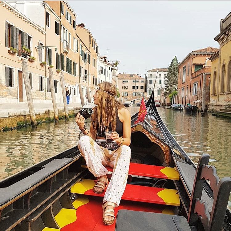 Woman in white Thai harem yoga pants with peacock print and black top in a gondola on a canal in Venice, Italy