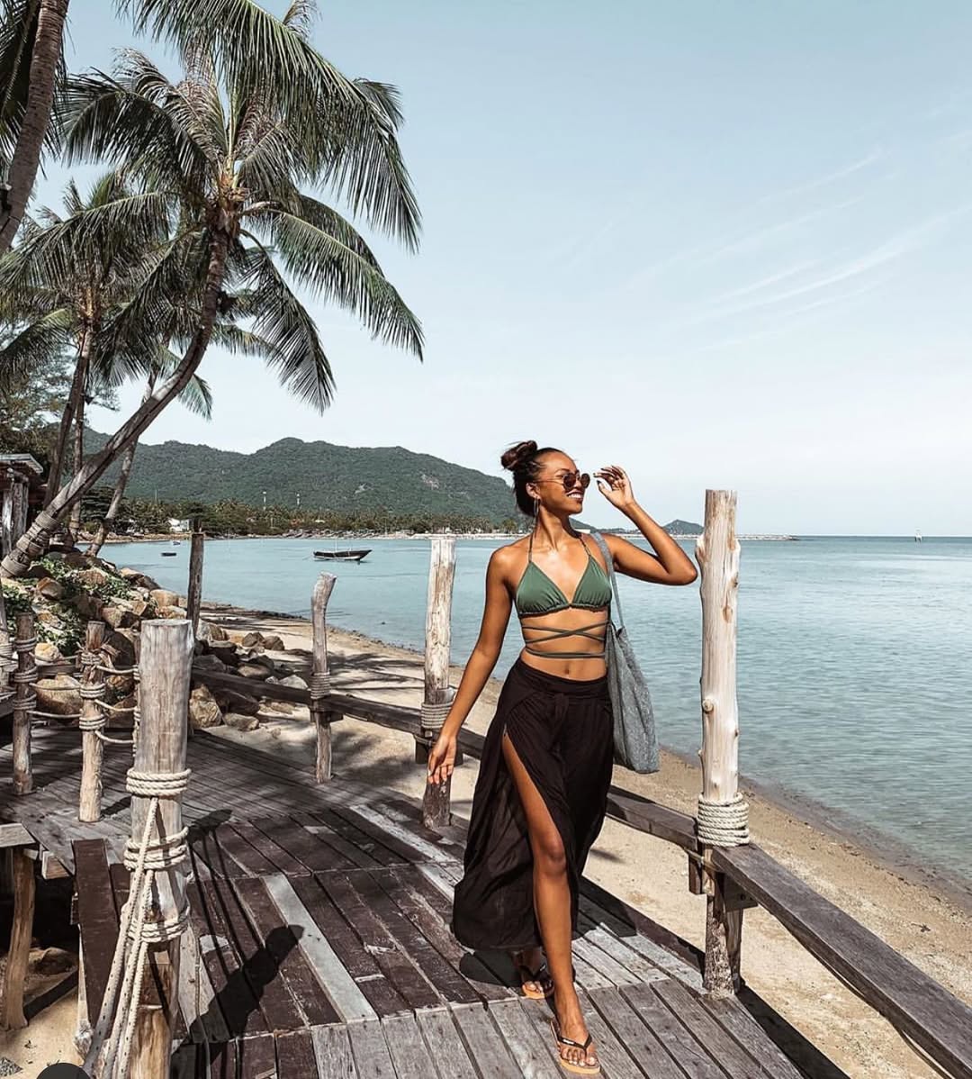 Woman in black Thai wrap pants skirt and green top standing on a wooden pier by the ocean with palm trees and mountains in the background.