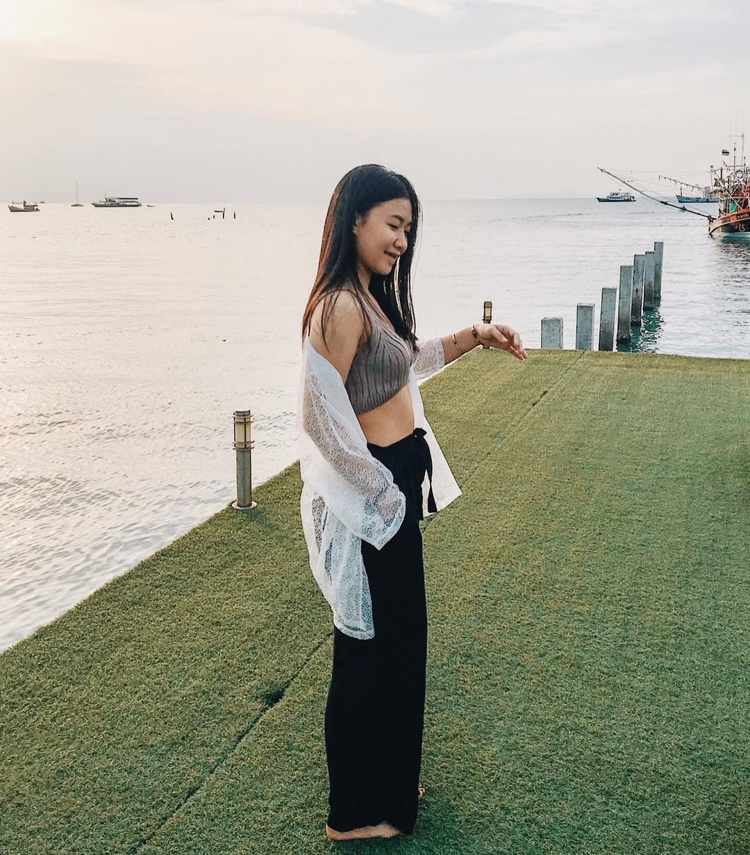 Woman standing in black Thai wrap pants skirt and grey top on a grassy area by a waterfront with boats in the background