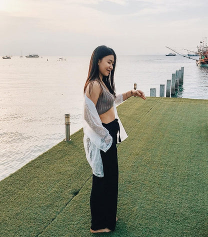 Woman standing in black Thai wrap pants skirt and grey top on a grassy area by a waterfront with boats in the background