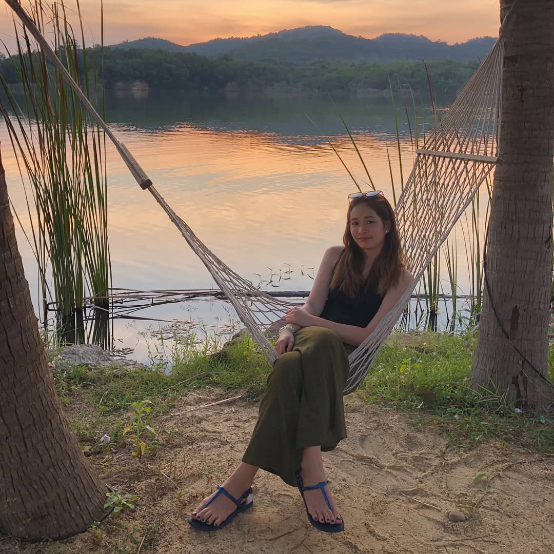 Woman sitting in a hammock by a lake at sunset wearing a green Thai wrap pants skirt and black top