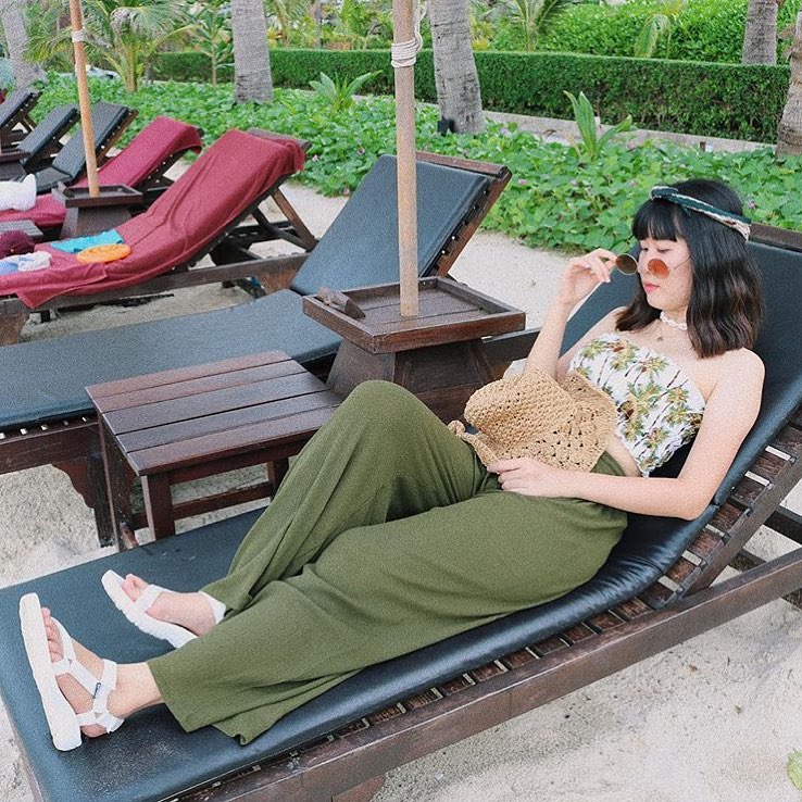 Woman in green Thai wrap pants skirt and beige top relaxing on a lounge chair in a tropical setting