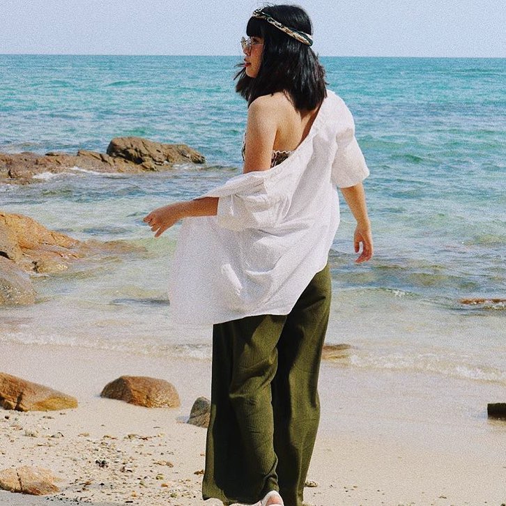 Woman in white shirt and green thai wrap pants skirt standing on a beach with clear blue water and rocks.