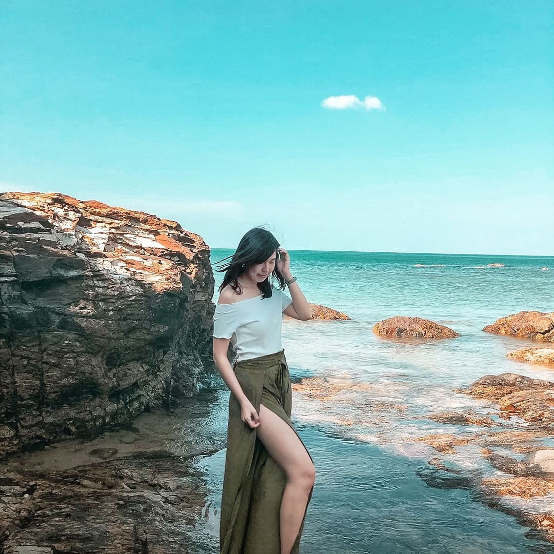 Woman  in green Thai wrap pants skirt and beige top standing on a rocky beach with clear blue water and sky.