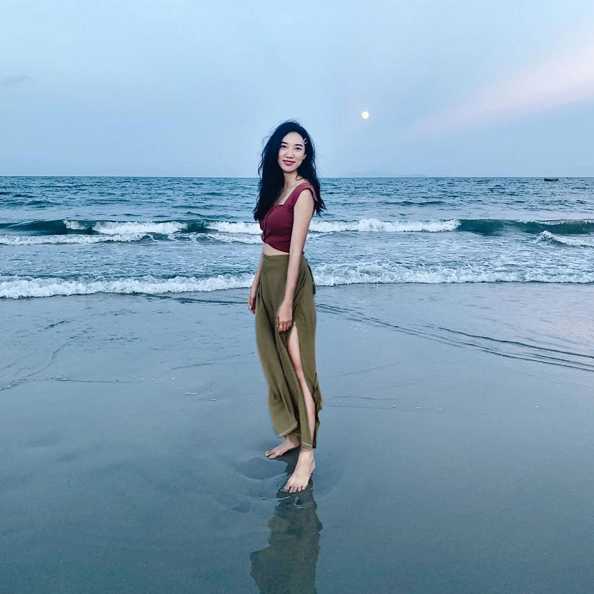 Woman in green Thai wrap pants skirt and purple top standing on a beach with ocean waves and a rainbow in the background