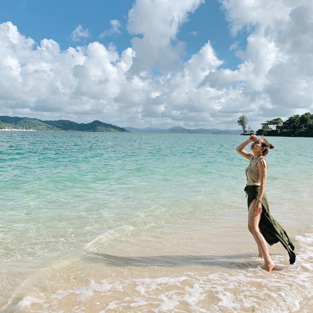 Woman in green Thai wrap pants skirt and beige top standing on a beach with clear turquoise water and blue sky.