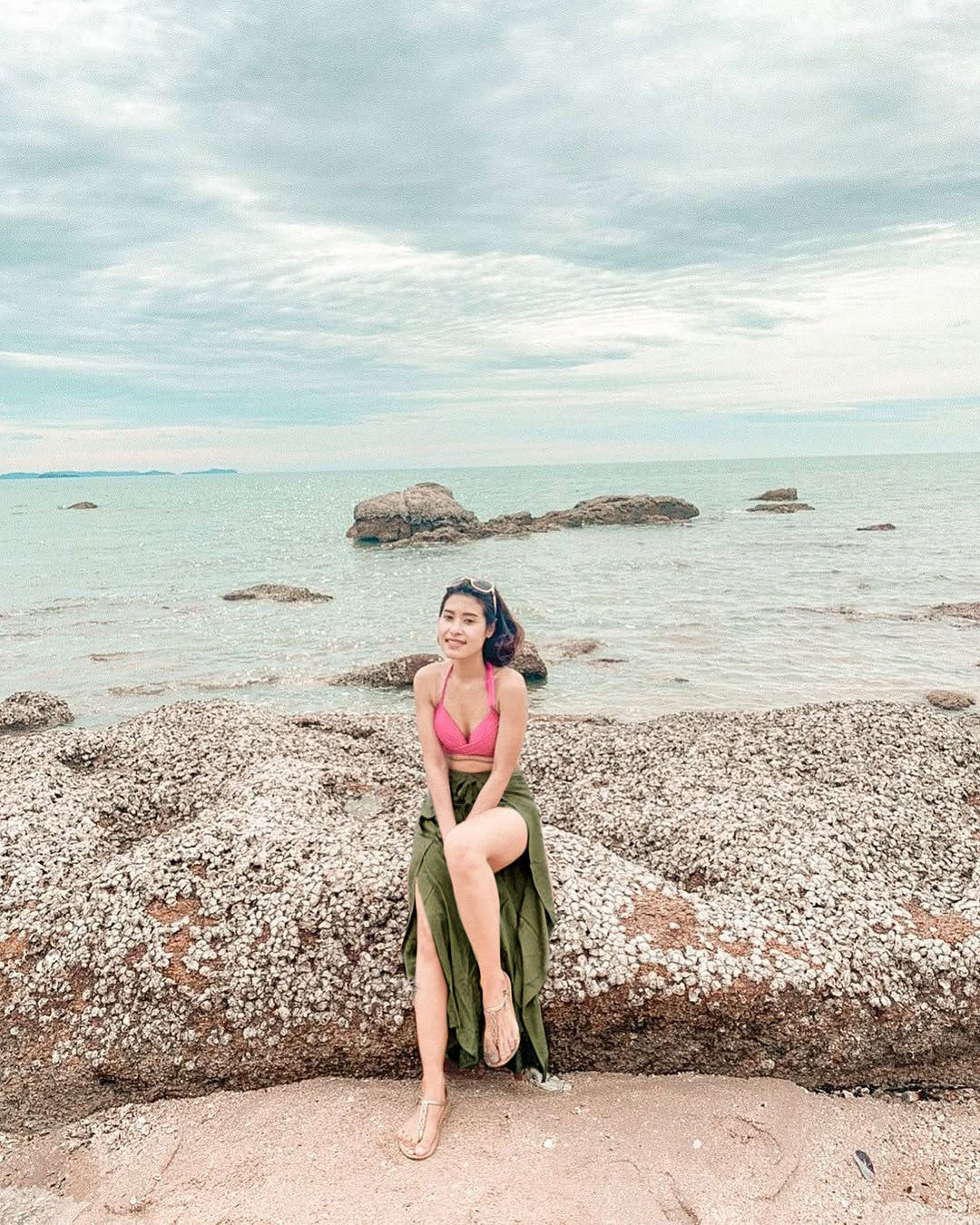 Woman in green Thai wrap pants skirt and pink top sitting on a rock by the sea with a cloudy sky.